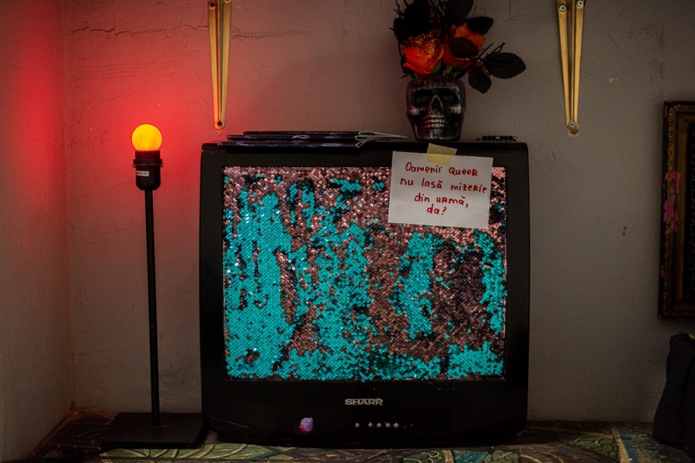 На фото старый телевизор Sharp. Вместо экрана панель обтянутая тканью с блестками. На панель приклеена кусок белой бумаги с красными буквами. На телевизоре черно-белая ваза в виде черепа с букетом из искусственных красных цветов. Также сверху стопкой лежат три блокнота. Слева стоит настольная лампа без абажура с ярко-желтой лампочкой.