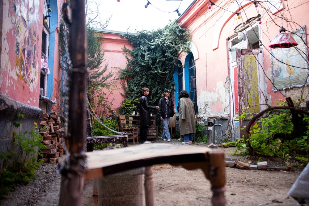 На фото внутренний двор с растениями, в него выходят три окна и дверь. Вверху висит незажженная гирлянда из разноцветных лампочек и абажур, на стене справа картина в раме. На переднем плане в расфокусе стул. В глубине двора у стола и стульев стоит трое людей. Один человек в сером плаще повернут спиной к камере. Два других человека в темной одежде разговаривают с этой персоной