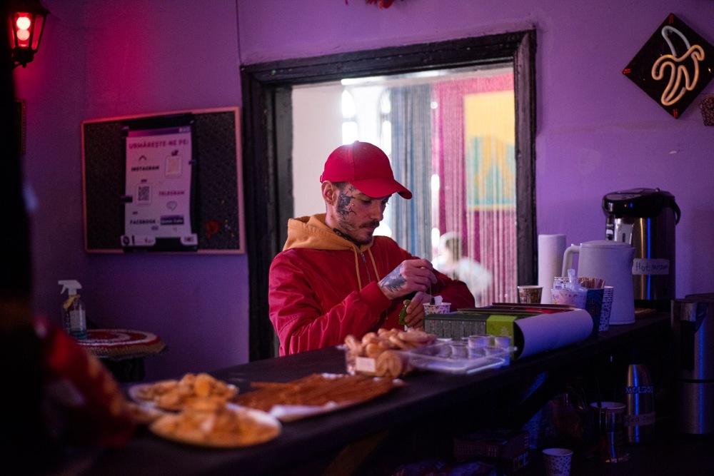 На фото комната с сиреневыми стенами. Человек в красно-желтой кофте и красной кепке с татуировками на лице и руках делает чай за барной стойкой. В расфокусе на стойке разная еда, чай, чайник и термос. На заднем плане проход в другую комнату.