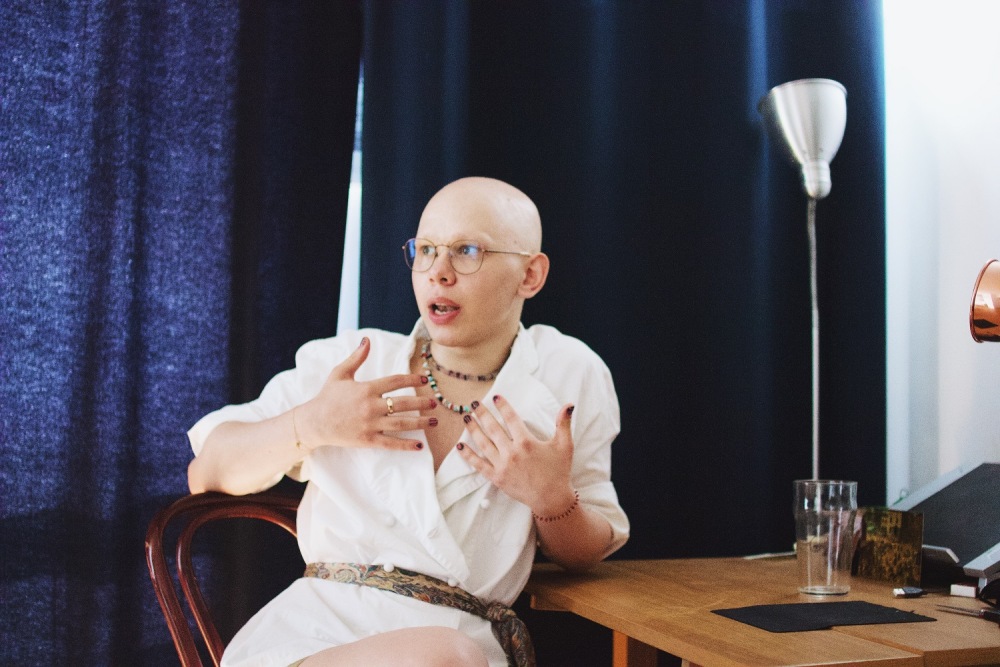 На фото Денис сидят на стуле в белом платье с разноцветным поясом, на них разноцветные бусы и два браслета на обеих руках, на среднем пальце правой руки кольцо. Он в очках, на голове нет волос, а на руках темно-красный маникюр. Они что-то рассказывают показывая двумя руками на себя и смотря вправо, облокотившись левым локтем на стол и закинув ногу за ногу. На столе стоят пустой стакан, лампа, кабели. На заднем плане темно-синие шторы.