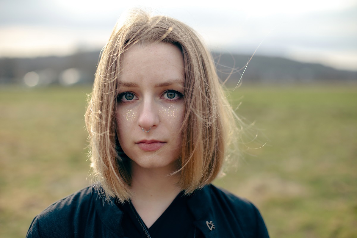 На фото Вера, у нее светлые прямые волосы до плеч, серо-голубые глаза. На щеках золотые блестки и пирсинг в септуме. Она смотрит в кадр, одета в черное.